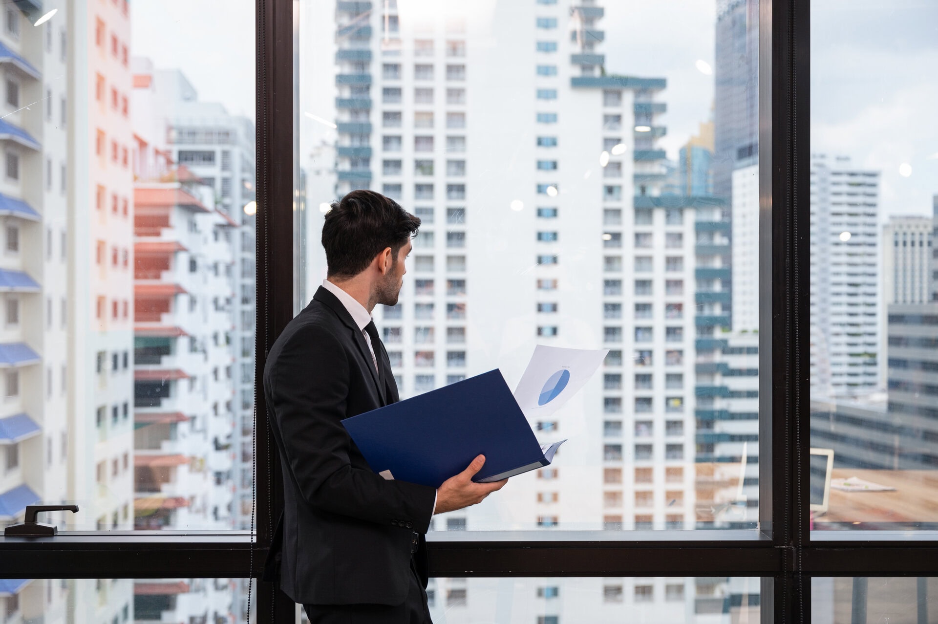 Modern businessman reviewing property documents in high-rise office with city skyline view.